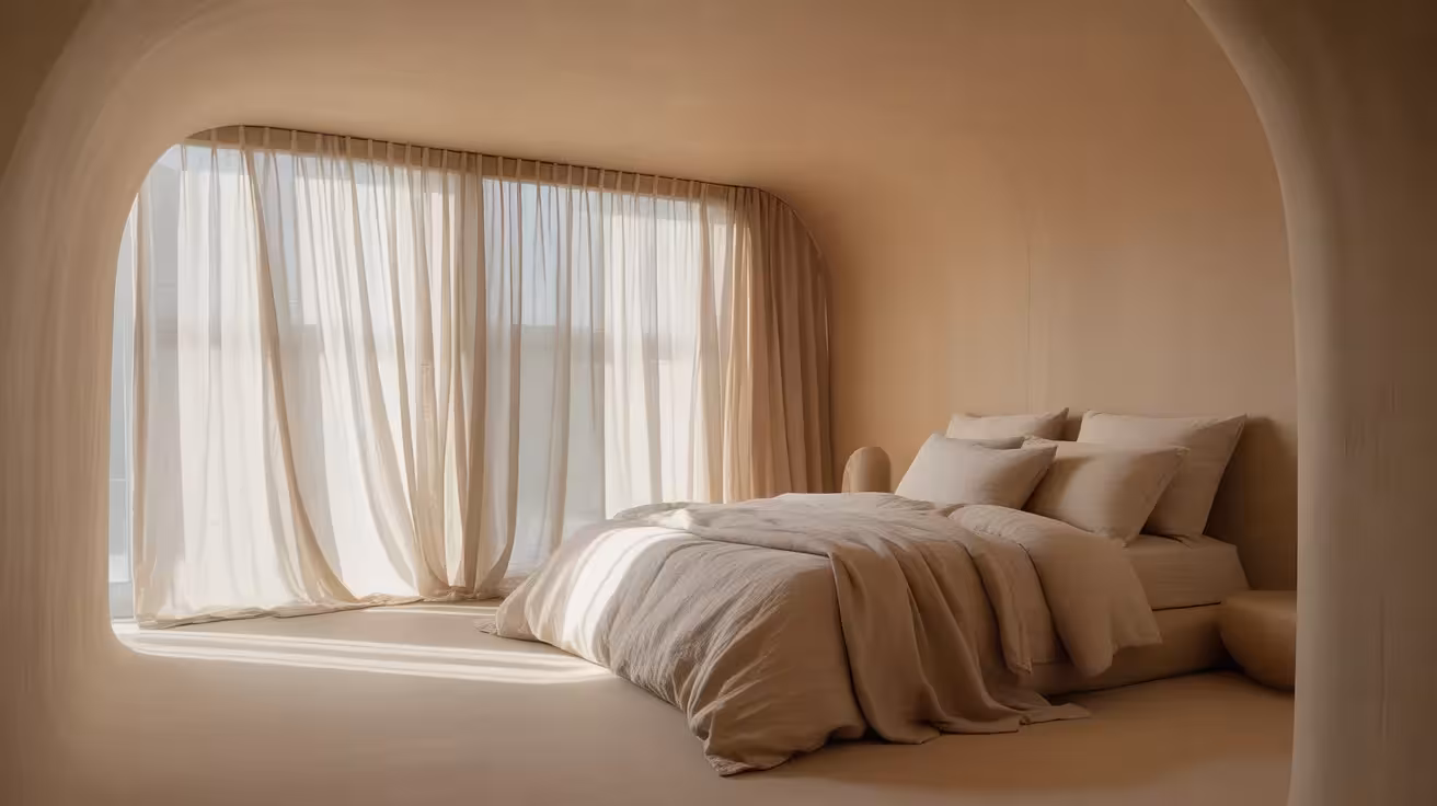 A wide view of a minimalist bedroom decorated in monolithic shades of warm beige, with soft morning light casting gentle shadows.