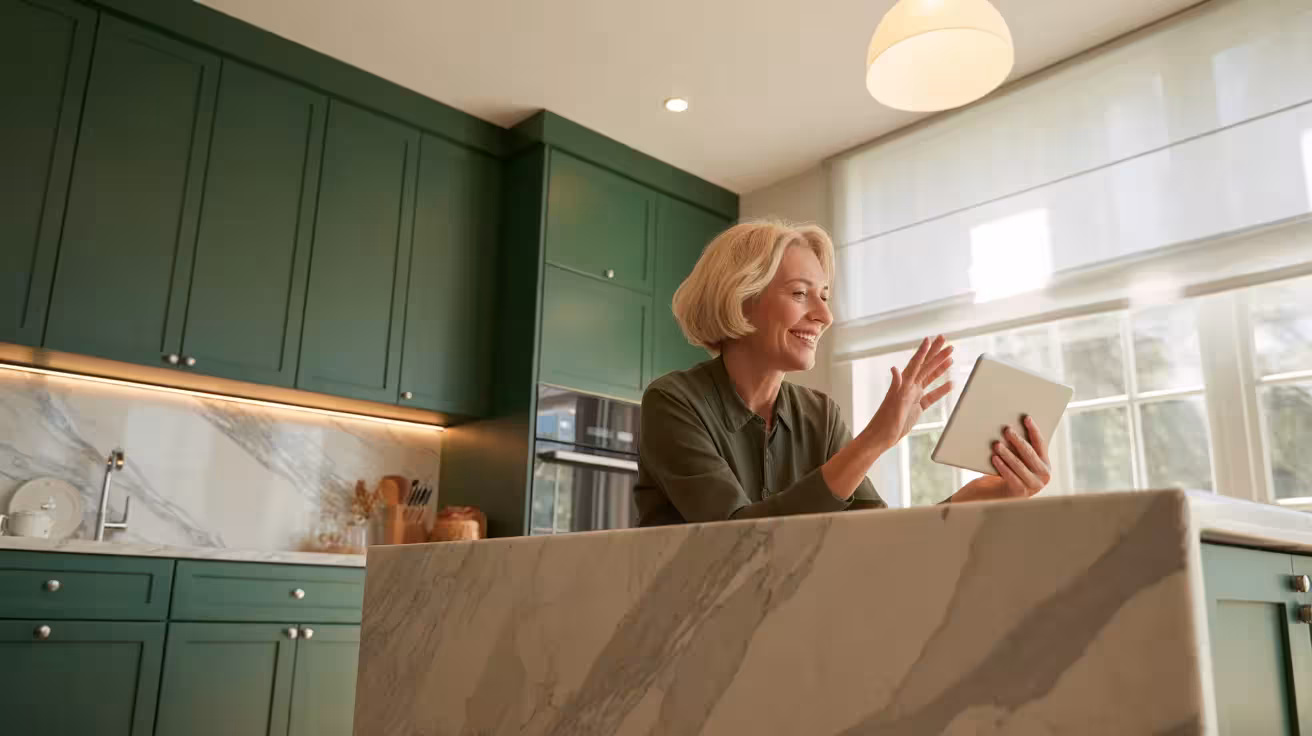 A senior woman on a video call in a spacious, modern green and marble kitchen, brightly backlit by a large window.
